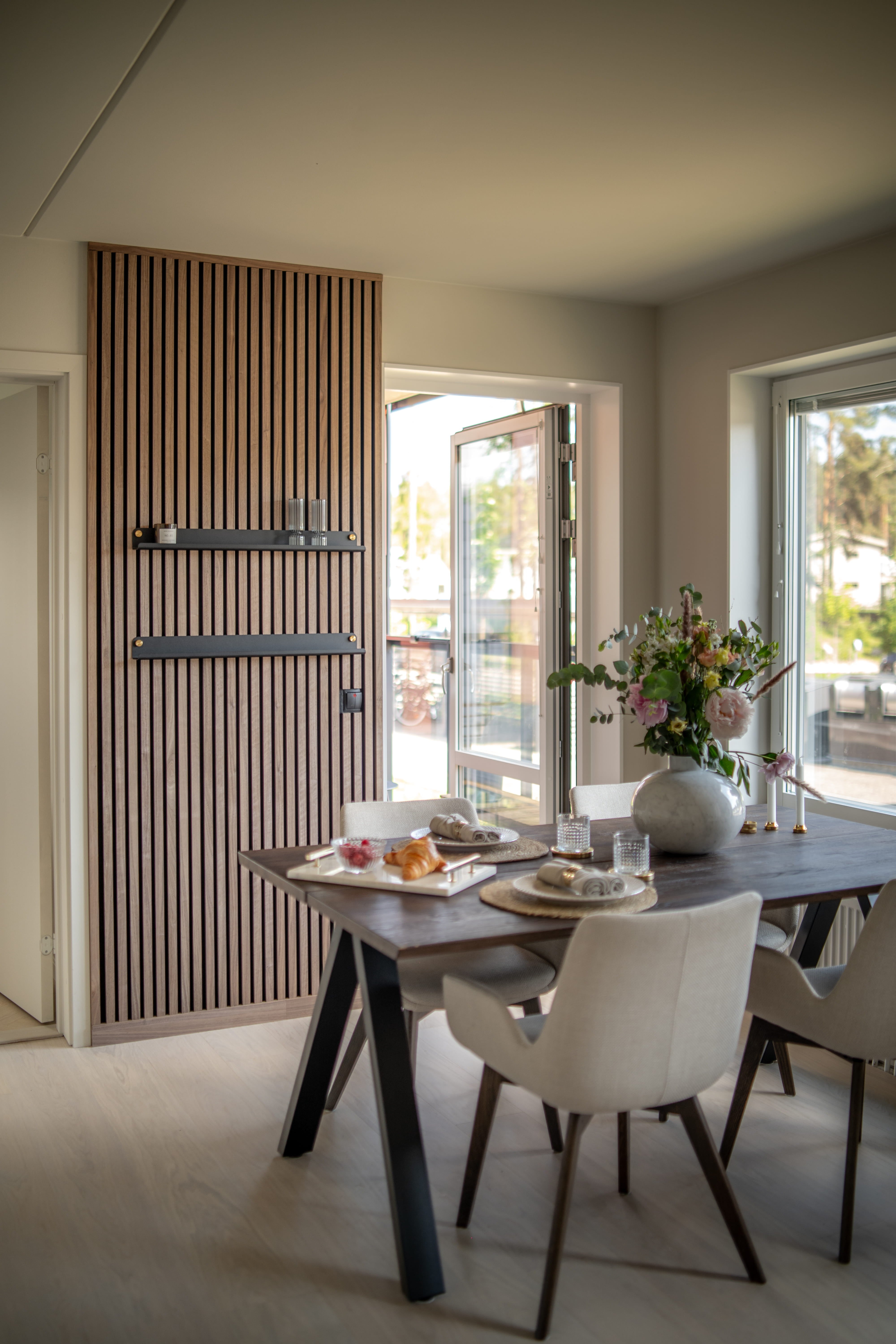 Modern matsal med texturerad Akupanel akustisk träribbvägg, mörkt frukostbord, minimalistiska beige stolar och en vas med blommor vid stora öppna fönster.